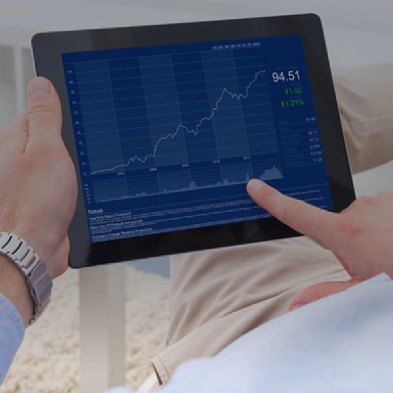 Person wearing beige pants and a light blue shirt, with a silver watch on the left hand, holding a tablet and pointing to stock data